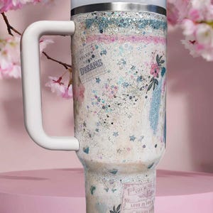 May include: A white insulated tumbler with a handle, lid, and straw. The tumbler is decorated with a glittery design, floral accents, and the phrases "Follow Your Dreams" and "Love is for Joy".
