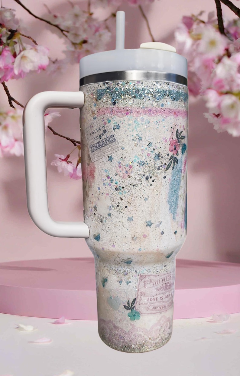 May include: A white insulated tumbler with a handle, lid, and straw. The tumbler is decorated with a glittery design, floral accents, and the phrases "Follow Your Dreams" and "Love is for Joy".