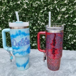 Puede incluir: Dos vasos de acero inoxidable con asa, tapa transparente y pajita. El vaso izquierdo tiene un patr&oacute;n de remolinos brillantes en azul claro y blanco con el nombre Debbie en azul. El vaso derecho tiene un acabado brillante en rojo y morado con el nombre Amy Dean en negro. Ambos tienen peque&ntilde;os dijes decorativos en las asas.