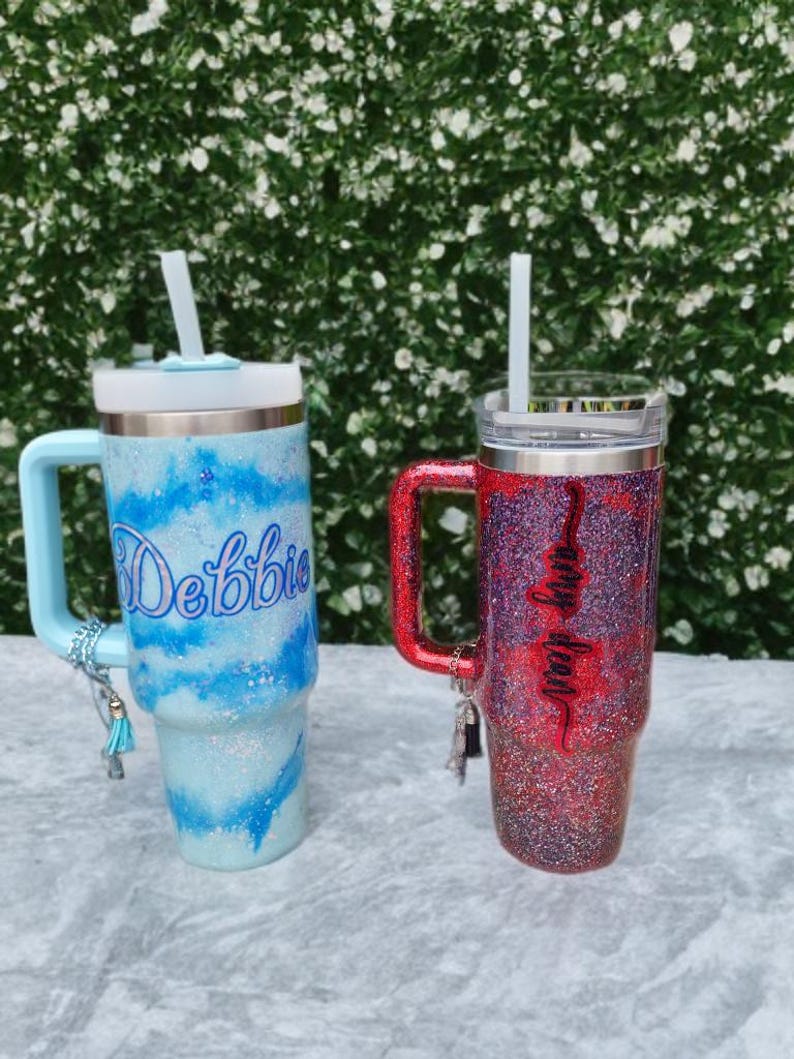 Puede incluir: Dos vasos de acero inoxidable con asa, tapa transparente y pajita. El vaso izquierdo tiene un patr&oacute;n de remolinos brillantes en azul claro y blanco con el nombre Debbie en azul. El vaso derecho tiene un acabado brillante en rojo y morado con el nombre Amy Dean en negro. Ambos tienen peque&ntilde;os dijes decorativos en las asas.