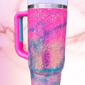 Puede incluir: Un vaso de acero inoxidable con asa, que presenta un acabado brillante en rosa y azul con un patr&oacute;n de remolinos tipo m&aacute;rmol. El vaso incluye una tapa blanca con un cierre deslizante rosa y un asa rosa con un acento central de color azul.