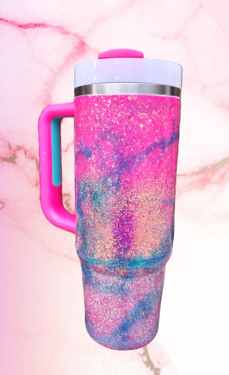 Puede incluir: Un vaso de acero inoxidable con asa, que presenta un acabado brillante en rosa y azul con un patr&oacute;n de remolinos tipo m&aacute;rmol. El vaso incluye una tapa blanca con un cierre deslizante rosa y un asa rosa con un acento central de color azul.