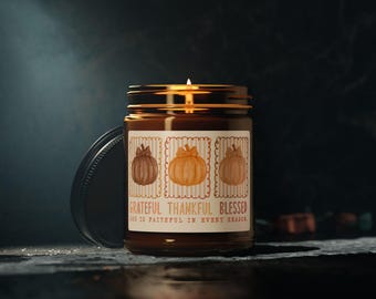 May include: A lit, brown glass jar candle with a black lid. The label features three pumpkin illustrations and the text "Grateful, Thankful, Blessed". The candle is on a dark surface.