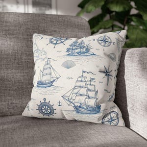 Könnte beinhalten: Quadratisches Kissen mit nautischem Thema. Das cremefarbene Kissen zeigt blaue Illustrationen von Segelbooten, Kompassen, Karten und anderen maritimen Elementen. Das Kissen liegt auf einem grauen Sofa.