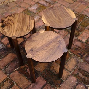 Puede incluir: Tres mesas auxiliares de madera con acabado en madera natural y detalles en latón. Las mesas están dispuestas en forma triangular sobre una superficie de ladrillo.