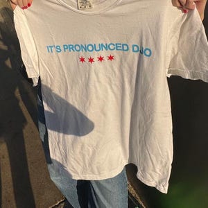 Op de afbeelding: Wit T-shirt met de tekst "IT'S PRONOUNCED DUO" in blauw en vier rode sterren. Het T-shirt wordt door een persoon omhoog gehouden. Het is een casual kledingstuk.