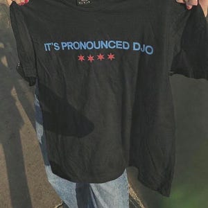 Könnte beinhalten: Schwarzes T-Shirt mit dem blauen Text "IT'S PRONOUNCED DJO" über vier roten Sternen. Das T-Shirt wird von einer Person hochgehalten. Es ist ein Freizeitkleidungsstück.