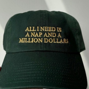 May include: A dark forest green baseball cap featuring the embroidered text ALL I NEED IS A NAP AND A MILLION DOLLARS in gold thread across the front panels. The cap has a structured crown, a curved brim, and visible stitching details.