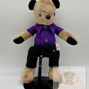 May include: A plush teddy bear with tan fur, black ears, and black paws. It wears a purple button-down shirt with a textured pattern and black pants with a drawstring. A white tag on the side reads Prima Dawna Bears, and a logo is visible.