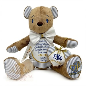 May include: A brown plush teddy bear with blue and white striped fabric on the ears, paws, and feet. It wears a white bow tie and holds a heart-shaped pillow. The bear features embroidered text and dates, including the name Phillip.