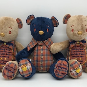 May include: Three plush teddy bears sitting in a row. The two outer bears are tan with plaid accents, embroidered text, and a heart with wings on their feet. The centre bear is dark blue with a plaid shirt featuring buttons and a collar.