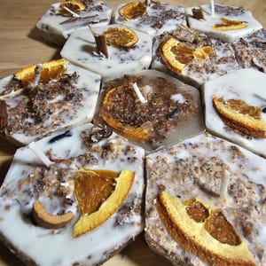 May include: A collection of hexagonal candles with a white base and a brown, speckled top. Each candle has a dried orange slice embedded in the wax and a small wick.