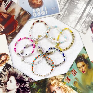Puede incluir: Varios brazaletes de cuentas de colores con palabras como "LOVER" y "FEARLESS". Portadas de álbumes con imágenes de una cantante están dispuestas alrededor de los brazaletes.