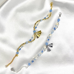 May include: Two beaded bracelets. One is gold-toned with blue and white beads, and a gold fan charm. The other is silver-toned with blue and white beads, and a silver fan charm. Both bracelets are on a white satin surface.