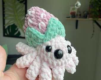May include: A handmade crochet octopus plushie in soft pink with a strawberry-like head. The head features pink and white sections with a green leaf-like trim. The octopus has black button eyes and eight tentacles, creating a whimsical and cuddly design.