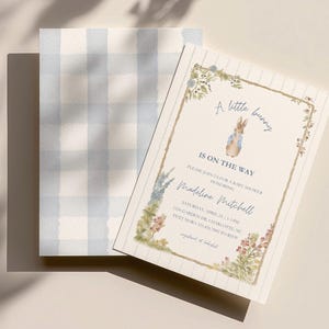 Può includere: Un invito per un baby shower con un'illustrazione di Peter Rabbit e dettagli floreali. Il testo dell'invito recita "A little bunny is on the way" con i dettagli dell'evento. Dietro l'invito è visibile una carta con un motivo a quadretti vichy blu e bianco.