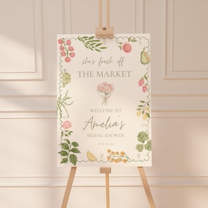 Puede incluir: Un letrero de bienvenida en un caballete de madera con el texto "she's fresh off THE MARKET" y "Welcome to Amelia's BRIDAL SHOWER". El letrero está decorado con ilustraciones de acuarela de frutas y flores.