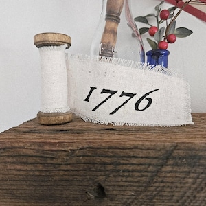 May include: A rustic display featuring a wooden spool wrapped with white fabric, a banner with the year "1776" in black, and a small blue vase with red berries. The items rest on a weathered wooden surface.