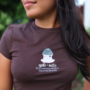 May include: A dark brown short-sleeve t-shirt featuring a graphic of a light blue and white creature wearing a shell hat. Below the graphic, the text reads: opihi-mistic the unwavering strength to cling on and persevere in white lettering.