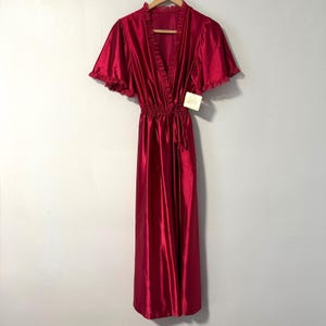 May include: A long, red satin robe with short sleeves and a V-neck. The robe has a gathered waist and a tie closure. The sleeves and neckline are trimmed with a delicate ruffle. A tag is attached to the robe.