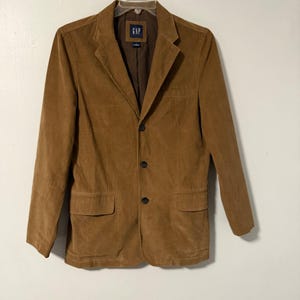May include: A brown corduroy blazer with a three-button closure and two front pockets. The blazer has a notched lapel and a small GAP label on the inside. The jacket is hanging on a white hanger.