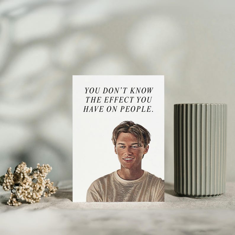 May include: A white greeting card with the text "YOU DON'T KNOW THE EFFECT YOU HAVE ON PEOPLE" and a portrait of a smiling person. A gray, ribbed vase is to the right of the card. A small bouquet of dried flowers is in the lower left corner.