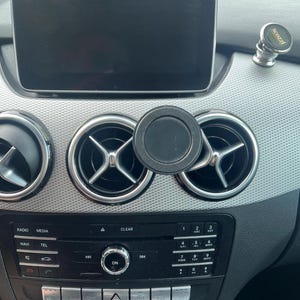 Op de afbeelding: Auto dashboard met een zwart infotainment scherm en ventilatieopeningen. Een zwarte magnetische telefoonhouder is bevestigd aan de ventilatieopening. Een zilveren en zwarte scozzi telefoonhouder staat op het dashboard.