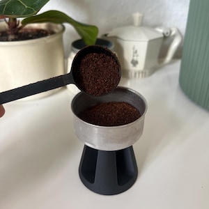 Könnte beinhalten: Ein schwarzer Kaffeelöffel aus Kunststoff, gefüllt mit dunkelbraunem Kaffeepulver, wird über einen Kaffeefiltereinsatz aus Metall gehalten. Der Filtereinsatz sitzt in einem schwarzen Kunststoffständer, der ebenfalls Kaffeepulver enthält.