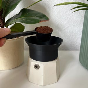 Könnte beinhalten: Ein schwarzer Löffel mit gemahlenem Kaffee wird in einen weiß-schwarzen Espressokocher gegossen. Der Espressokocher hat einen silbernen Schalter. Grüne Pflanzen in Töpfen sind im Hintergrund.