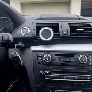 May include: A car dashboard with a silver and black phone mount attached to the air vent. The dashboard features various buttons, dials, and a start/stop button. A car key is hanging from the ignition.