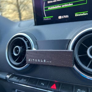 Könnte beinhalten: Ein dunkelbrauner Holz-Autolüftungsclip mit dem Wort "RITUALS..." in weißer Schrift. Der Clip ist an einem Armaturenbrett in der Nähe der Lüftungsöffnung befestigt. Der Infotainment-Bildschirm des Autos ist im Hintergrund sichtbar.