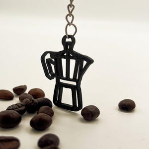 Könnte beinhalten: Ein schwarzer Metall-Schlüsselanhänger mit einem Kaffeemaschinen-Design, der an einer Kette hängt. Mehrere geröstete Kaffeebohnen sind um den Schlüsselanhänger auf einer weißen Oberfläche verstreut. Ein stilvolles Accessoire für Kaffeeliebhaber.