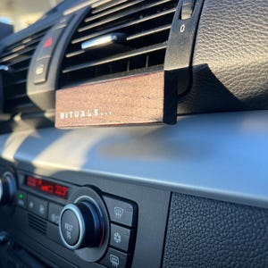 Può includere: Diffusore per bocchetta dell'aria dell'auto in legno marrone con la scritta "RITUALS...". Il diffusore è rettangolare e si inserisce nella bocchetta dell'aria dell'auto. Il cruscotto dell'auto è nero e argento.