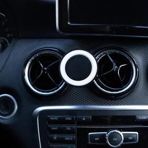 May include: A black and white car vent phone mount is attached to the dashboard. The mount has a circular design with a white outer ring and a black center. The car's dashboard is black with silver accents.