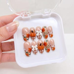 May include: A set of ten press-on nails arranged in a white plastic case. The nails feature a nude base with a brown and white gingham pattern at the tips, accented with 3D orange pumpkins, white bows, and small red and orange autumn leaves.