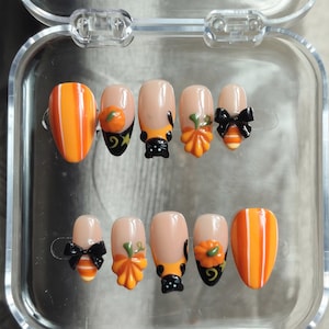 Peut inclure: Un ensemble de dix faux ongles dans un boîtier en plastique transparent, avec une finition brillante, des rayures verticales orange et blanches, des nœuds noirs, des citrouilles orange et des motifs de chats noirs sur une base nude.
