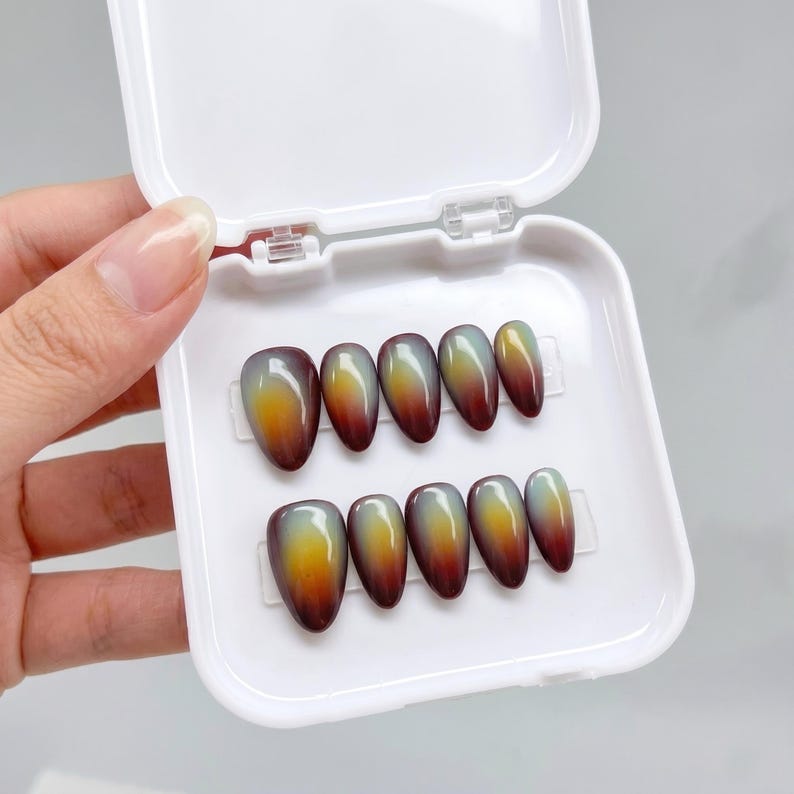May include: A set of ten almond-shaped press-on nails arranged in a white plastic storage case. Each nail features a smooth, glossy gradient finish that transitions from a deep burgundy at the tip to a vibrant yellow and light green at the base.