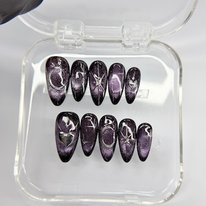 May include: A set of ten almond-shaped press-on nails in a deep purple magnetic cat-eye finish. Each nail features silver hand-painted celestial designs including crescent moons, four-pointed stars, and hearts, arranged in a clear plastic storage case.