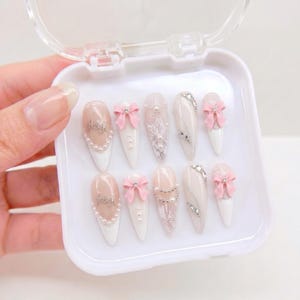 May include: A set of ten almond-shaped press-on nails in a white plastic case. The nails feature a white and nude color palette with pink 3D bows, silver lettering, white lace patterns, small pearls, and clear rhinestones arranged in various designs.