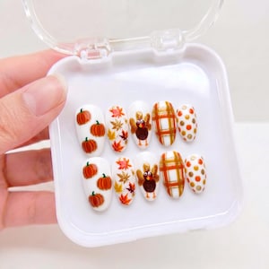 May include: A set of ten white press-on nails arranged in a white plastic case. The designs feature orange pumpkins, multicolored autumn leaves, brown turkeys, a gold and orange plaid pattern, and orange and gold polka dots on each nail.