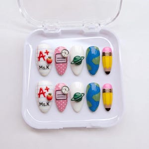 May include: A set of ten press-on nails in a white plastic case. Designs include lined paper with A+ and Ms.K, pink polka dots with a clock, white with a 3D planet, blue and green earth patterns, and yellow pencils with pink erasers.