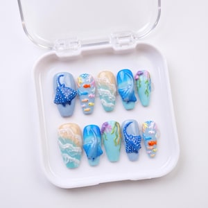 以下が含まれることがあります： 白いプラスチックケースに入った10枚のネイルチップセット。青いエイ、カラフルな小魚、白い波、緑の海藻などの3Dデザインが、水色や砂色のベースに施されています。