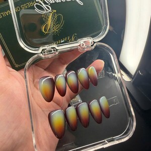 May include: A set of ten almond-shaped press-on nails featuring a gradient color transition from a deep dark brown at the tips to a bright yellow and light green center, housed in a clear plastic storage case with a dark green branded insert.