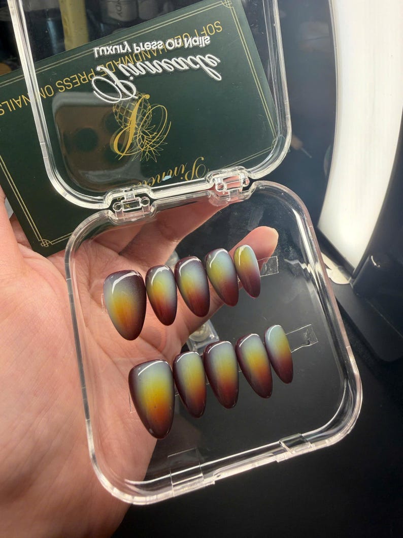 May include: A set of ten almond-shaped press-on nails featuring a gradient color transition from a deep dark brown at the tips to a bright yellow and light green center, housed in a clear plastic storage case with a dark green branded insert.