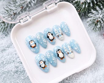 May include: A set of ten almond-shaped press-on nails in a light blue base color. The designs feature hand-painted penguins, white snowflakes, and snowy pine tree motifs. The nails are arranged inside a small, square, white plastic storage case.