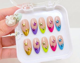 May include: A set of ten almond-shaped press-on nails in a white plastic case. Each nail features a nude base with a colorful French tip in yellow, purple, lime green, hot pink, or blue, accented with two small metallic star-shaped charms.