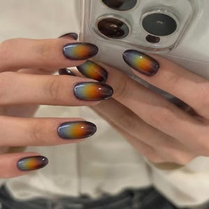 May include: Oval-shaped fingernails featuring a smooth gradient design that transitions from a dark charcoal gray at the tips to a vibrant orange and yellow center, fading into a soft light gray base near the cuticle, held against a smartphone.