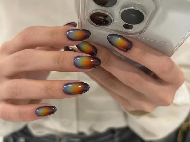 May include: Oval-shaped fingernails featuring a smooth gradient design that transitions from a dark charcoal gray at the tips to a vibrant orange and yellow center, fading into a soft light gray base near the cuticle, held against a smartphone.