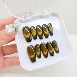 May include: A set of ten press-on nails with a dark olive green cat-eye magnetic finish, arranged in a white plastic storage case. Each nail features a glossy, reflective band of light that shifts across the surface, creating a deep, dimensional effect.