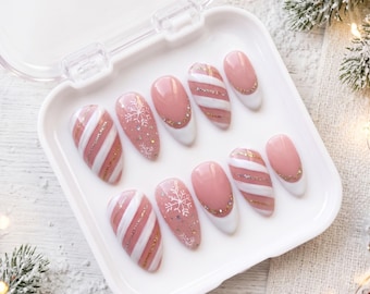 May include: A set of ten almond-shaped press-on nails in a white plastic case. The designs feature a nude base with white and gold glitter diagonal stripes, white snowflake patterns, and nude nails with white tips and gold glitter accents.